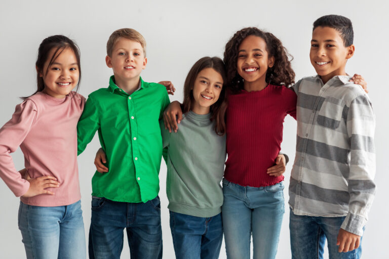 Cheerful Multicultural Preteen Kids Standing Hugging And Smiling To Camera Posing Together Over Gray Studio Background. Shot Of Mixed School Boys And Girls Group. Friendship Concept