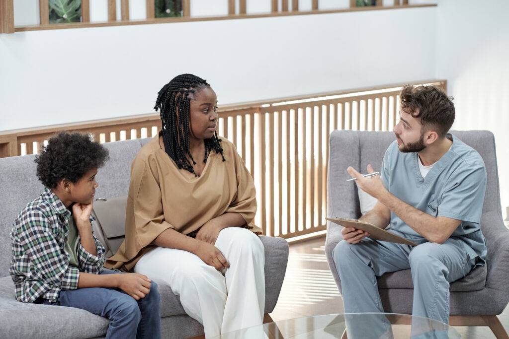 Young confident dentist giving consultation to young African woman with son while sitting in lounge of dental clinics